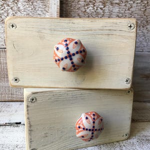 May include: Two stacked, off-white painted wooden boxes with decorative ceramic knobs. The knobs are orange, white, and blue, featuring a floral design. The boxes are held together with silver screws, showcasing a rustic, handcrafted aesthetic.