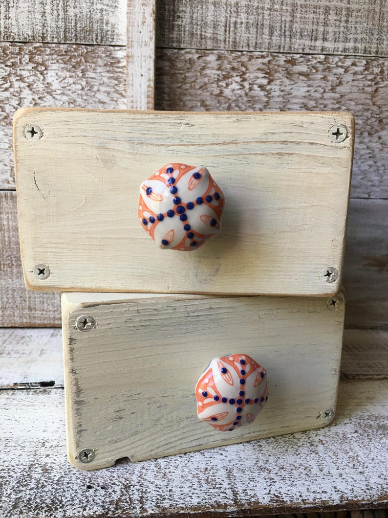 May include: Two stacked, off-white painted wooden boxes with decorative ceramic knobs. The knobs are orange, white, and blue, featuring a floral design. The boxes are held together with silver screws, showcasing a rustic, handcrafted aesthetic.