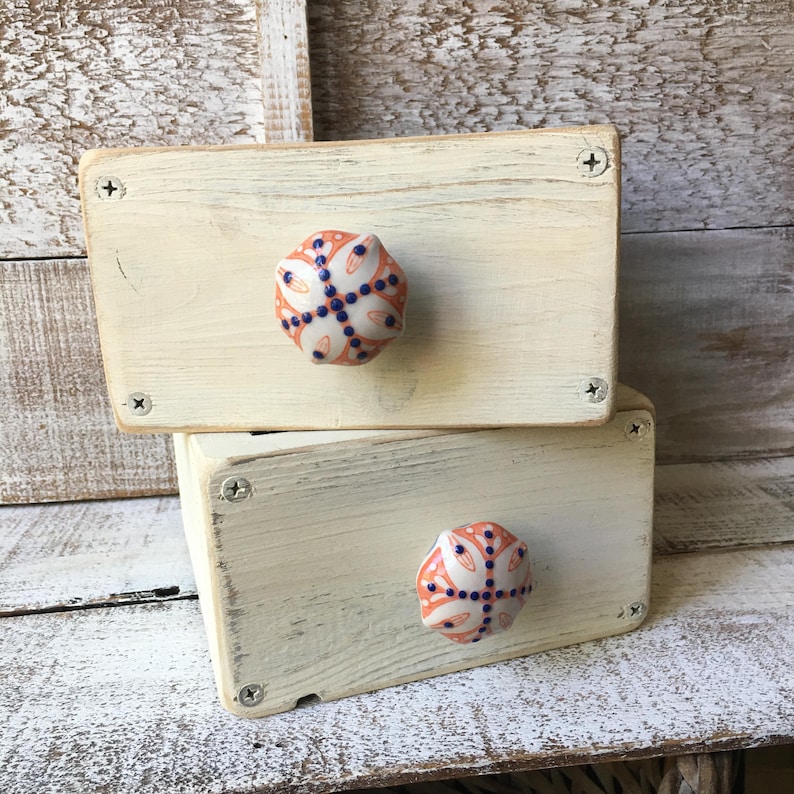 May include: Two small, off-white wooden boxes stacked on top of each other. Each box has a decorative ceramic knob with a floral design in coral, white, and navy blue. The boxes are held together with screws.