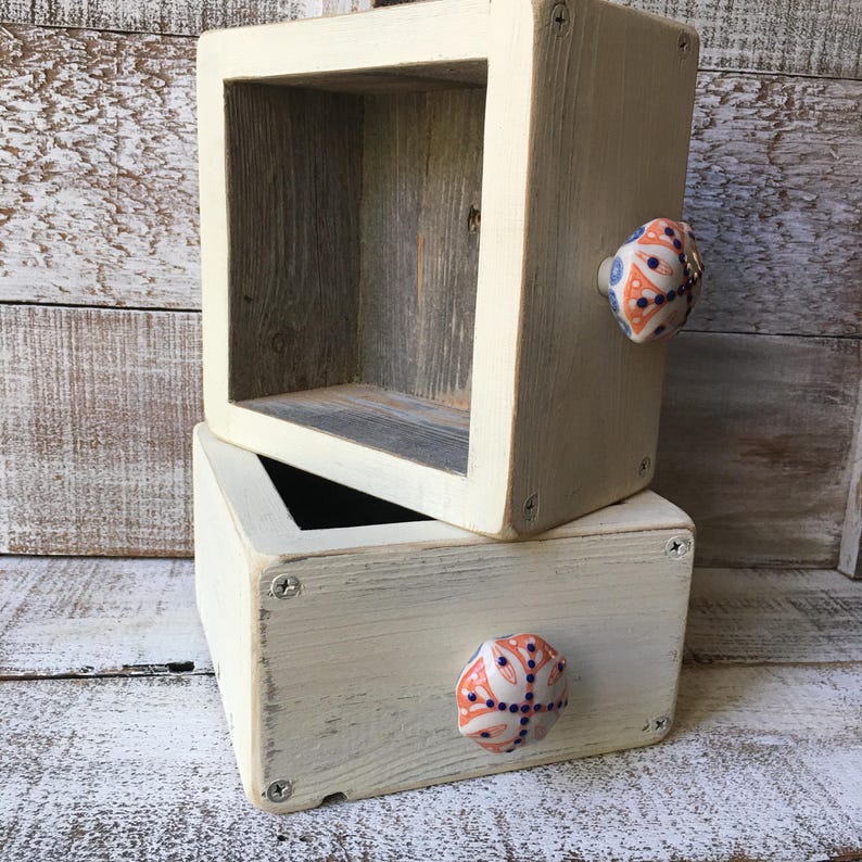 May include: Two stacked, square, wooden shelves with a distressed, off-white finish. Each shelf has a decorative knob with a white, orange, and blue floral design. The interior of the shelves is a natural wood color.