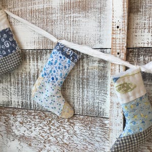 May include: A garland of three small, fabric Christmas stockings. Each stocking has a different pattern, including floral, paisley, and plaid designs. They are suspended from a white ribbon against a weathered, white-painted wooden background.