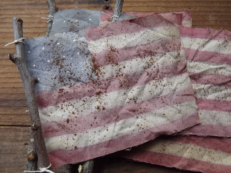 May include: Three American flags made of fabric with a distressed, vintage look. The flags are attached to sticks and have a faded red, white, and blue color scheme.