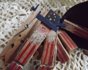 May include: Four wooden clothespins painted with red and white stripes and a blue and white hat. The clothespins are arranged to look like a person.