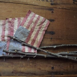 May include: Two small American flags made of fabric are tied together with twine and attached to a wooden crate with twigs. The flags are faded and have a rustic look.