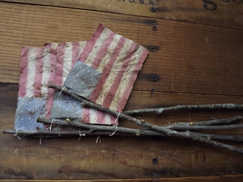 May include: Two small American flags made of fabric are tied together with twine and attached to a wooden crate with twigs. The flags are faded and have a rustic look.