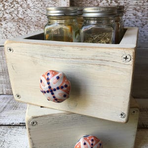 May include: A rustic, off-white wooden spice rack with two drawers, each adorned with a decorative ceramic knob featuring an orange and blue floral design. The top drawer holds several glass jars with silver lids, filled with herbs and spices.
