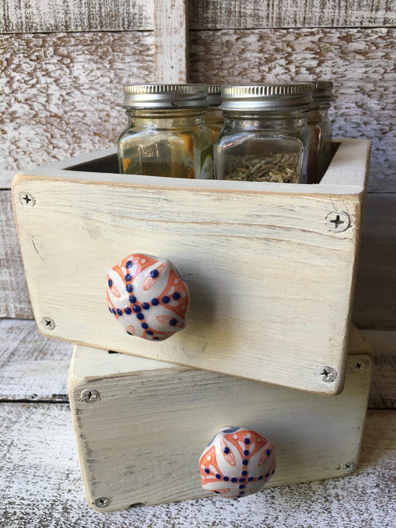 May include: A rustic, off-white wooden spice rack with two drawers, each adorned with a decorative ceramic knob featuring an orange and blue floral design. The top drawer holds several glass jars with silver lids, filled with herbs and spices.