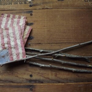 May include: A weathered wooden crate with a small American flag made of fabric and several thin branches laying across the top.
