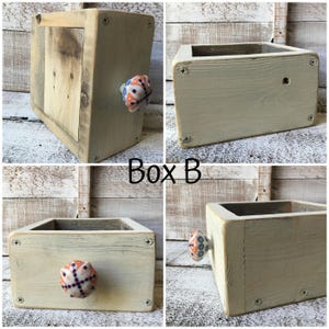 May include: A set of four wooden boxes, each with a rectangular shape and a light beige finish. One box features a decorative knob with blue, orange, and white patterns. The text "Box B" is visible.
