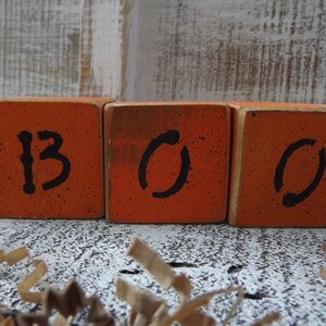 May include: Three orange wooden blocks with black painted letters spelling "BOO".