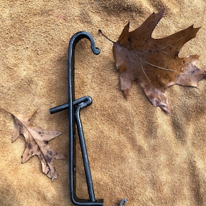 May include: Black metal hook with a unique design, featuring two curved hooks and a small bar. The hook is lying on a brown suede surface.