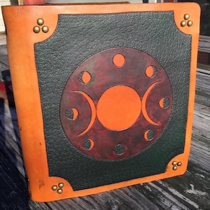May include: A square leather journal with a tan border and a textured dark green center panel. The cover features a central circular design with moon phases in shades of orange and deep red, accented by brass-colored metal studs at each corner.