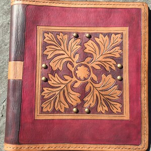 Könnte beinhalten: Ein quadratischer Einband aus Leder mit einem tief burgunderrot gefärbten Mittelteil, einem hellbraunen geprägten Blattmuster und einem zentralen Blumenmotiv. Der Einband ist mit kleinen Messingnieten verziert und hat einen hellbraunen Rand.