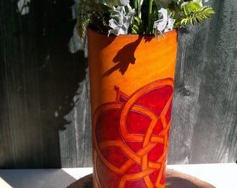 May include: Tall, cylindrical leather vase in shades of orange and red, adorned with a Celtic knot design. The vase is filled with greenery and rests on a wooden surface. The vase is approximately 12 inches tall.