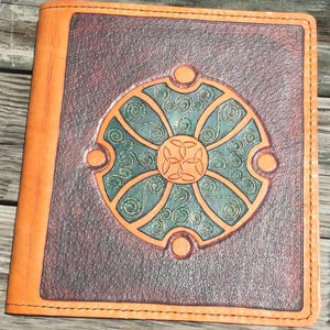 May include: A rectangular leather journal cover featuring a dark brown textured surface with a central circular emblem. The emblem displays a teal green background with intricate embossed spiral patterns and a central triquetra knot design.