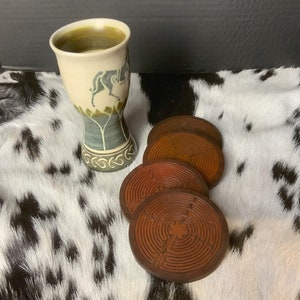 May include: Four brown coasters with a circular design, stacked on top of each other. The coasters are made of wood and have a natural finish. A ceramic mug with a green and white design is in the background.