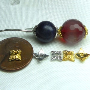 May include: A close-up of a jewelry making kit with two round beads, one blue and one red, and four gold and silver star-shaped bead caps. The beads are about the size of a US penny.