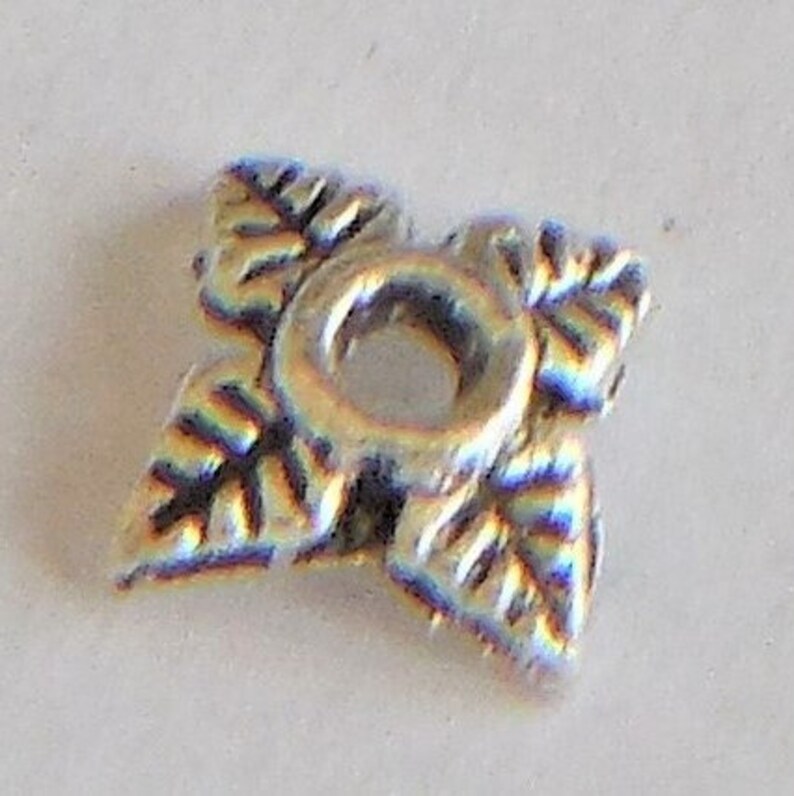 May include: Silver-toned metal bead with a decorative leaf design. The bead has a hole in the center for stringing.