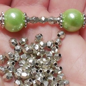 Op de afbeelding: Een close-up van een zilveren en groene kralenketting met twee grote groene kralen aan beide uiteinden. De ketting is omringd door een stapel kleine zilveren kralen.