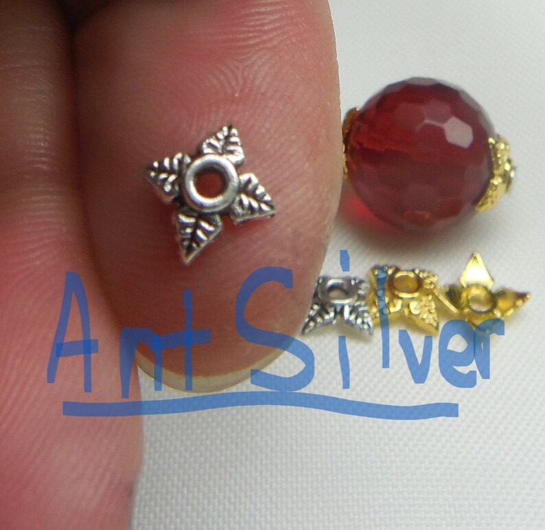 May include: Silver and gold tone metal bead caps with a star-shaped design. The silver cap has a round hole in the center. The gold cap has a leaf design.