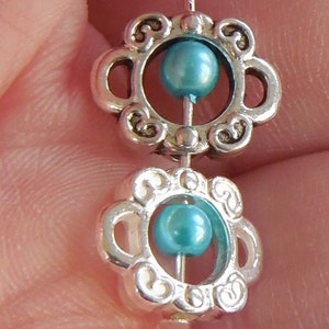 May include: Two silver charm beads with a teal blue glass bead in the center of each. The beads have a decorative floral design.