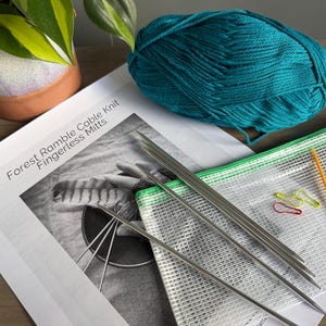 May include: A knitting kit featuring a teal yarn skein, a set of silver metal knitting needles, a clear mesh zipper pouch, two colorful stitch markers, and a printed pattern sheet titled Forest Ramble Cable Knit Fingerless Mitts.