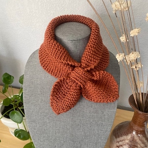 May include: A rust-colored knitted scarf with a bow tie design. The scarf is displayed on a gray mannequin.