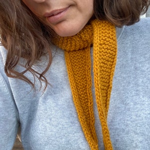 May include: A mustard yellow knitted scarf featuring a garter stitch texture and a tapered, pointed end design. The scarf is wrapped around the neck and worn over a light grey crew neck sweater, showcasing a simple, functional knit construction.