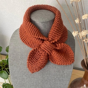 Könnte beinhalten: Ein handgestrickter Schal in Rostbraun, präsentiert auf einem grauen Mannequinkopf. Der Schal besteht aus dickem Garn und hat ein geschlungenes Design, das vorne gebunden ist. Die Textur des Strickwerks ist deutlich sichtbar.