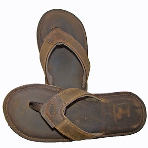 May include: A pair of brown leather flip-flops with a simple design. The flip-flops have a brown leather strap and a brown leather sole.