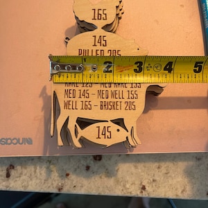 May include: Wooden cutouts of a chicken, pig, and fish with different internal temperature readings for cooking. The chicken is 165 degrees Fahrenheit, the pig is 145 degrees Fahrenheit, and the fish is 145 degrees Fahrenheit. The text "MED 145 - MED WELL 155 WELL 165 - BRISKET 205" is also included.