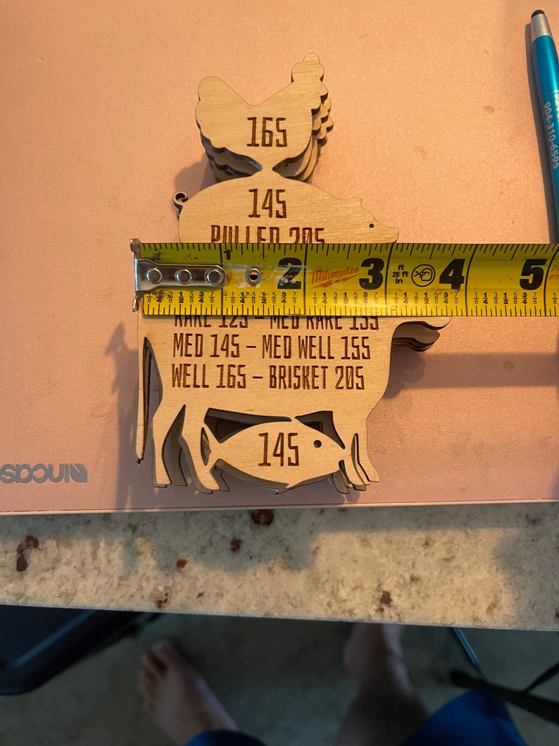 May include: Wooden cutouts of a chicken, pig, and fish with different internal temperature readings for cooking. The chicken is 165 degrees Fahrenheit, the pig is 145 degrees Fahrenheit, and the fish is 145 degrees Fahrenheit. The text "MED 145 - MED WELL 155 WELL 165 - BRISKET 205" is also included.