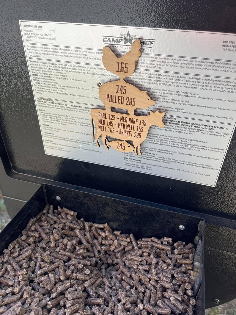 May include: A wooden grilling temperature guide with a chicken, pig, cow, and fish silhouette. The guide shows recommended temperatures for different types of meat, including rare, medium rare, medium, medium well, well, and brisket. The text on the guide reads "Camp Chef, The Way to Cook Outdoors, 145 Pulled 205, Rare 125 - Med Rare 135, Med 145 - Med Well 155, Well 165 - Brisket 205, 145".