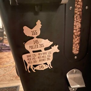 May include: A black metal smoker with a woodwind logo and a brown wood pellet hopper. A metal sticker with a chicken, pig, cow, and fish silhouette shows recommended cooking temperatures for different meats. The sticker reads "165", "145", "PULLED 205", "RARE 125 - MED RARE 135", "MED 145 - MED WELL 155", "WELL 165 - BRISKET 205", and "145".