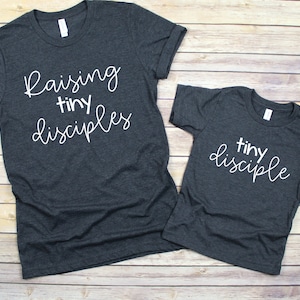 May include: Two grey t-shirts with white text. The larger shirt says "Raising tiny disciples". The smaller shirt says "tiny disciple".