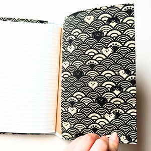Puede incluir: Cuaderno abierto con p&aacute;ginas rayadas. La cubierta presenta un patr&oacute;n en blanco y negro de olas estilizadas y caras de gatos. El cuaderno se sostiene abierto, revelando las p&aacute;ginas en blanco.