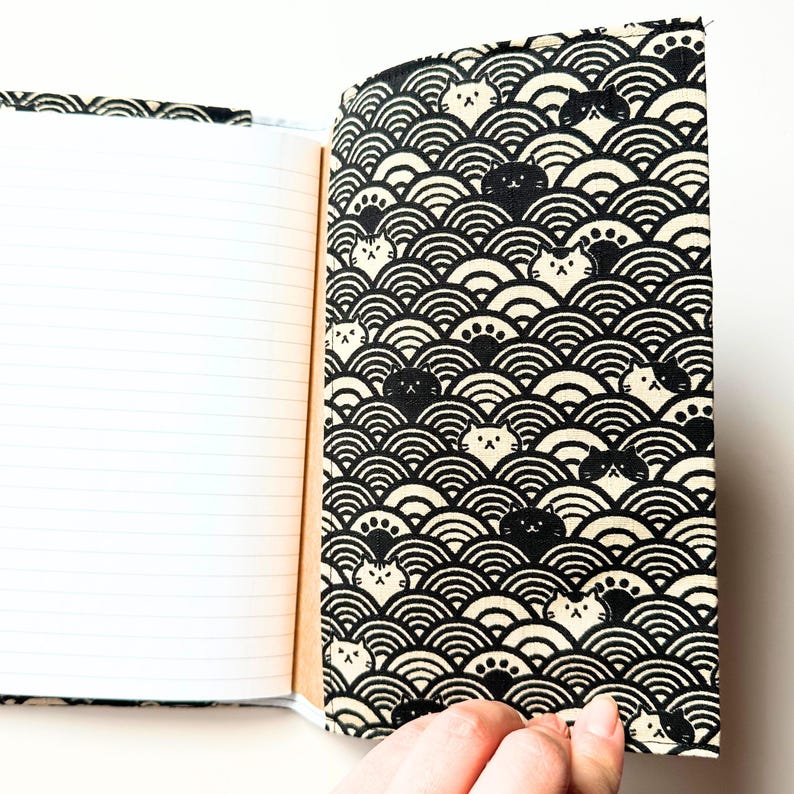 Puede incluir: Cuaderno abierto con p&aacute;ginas rayadas. La cubierta presenta un patr&oacute;n en blanco y negro de olas estilizadas y caras de gatos. El cuaderno se sostiene abierto, revelando las p&aacute;ginas en blanco.