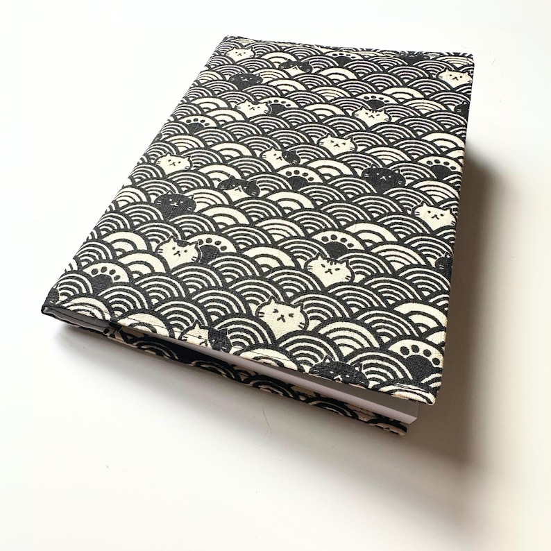 Puede incluir: Un cuaderno cerrado con un patr&oacute;n de olas en blanco y negro y caras de gato. La cubierta presenta un dise&ntilde;o repetitivo de olas negras y caras de gato blancas. El cuaderno es rectangular y parece estar hecho de tela.
