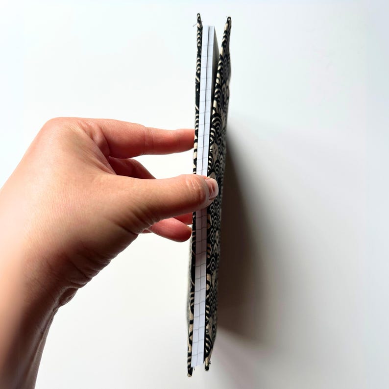 Puede incluir: Un cuaderno estampado con un dise&ntilde;o en negro y beige. El cuaderno est&aacute; abierto, revelando p&aacute;ginas rayadas. La cubierta tiene un patr&oacute;n de remolino repetido. El cuaderno se sostiene sobre un fondo blanco.
