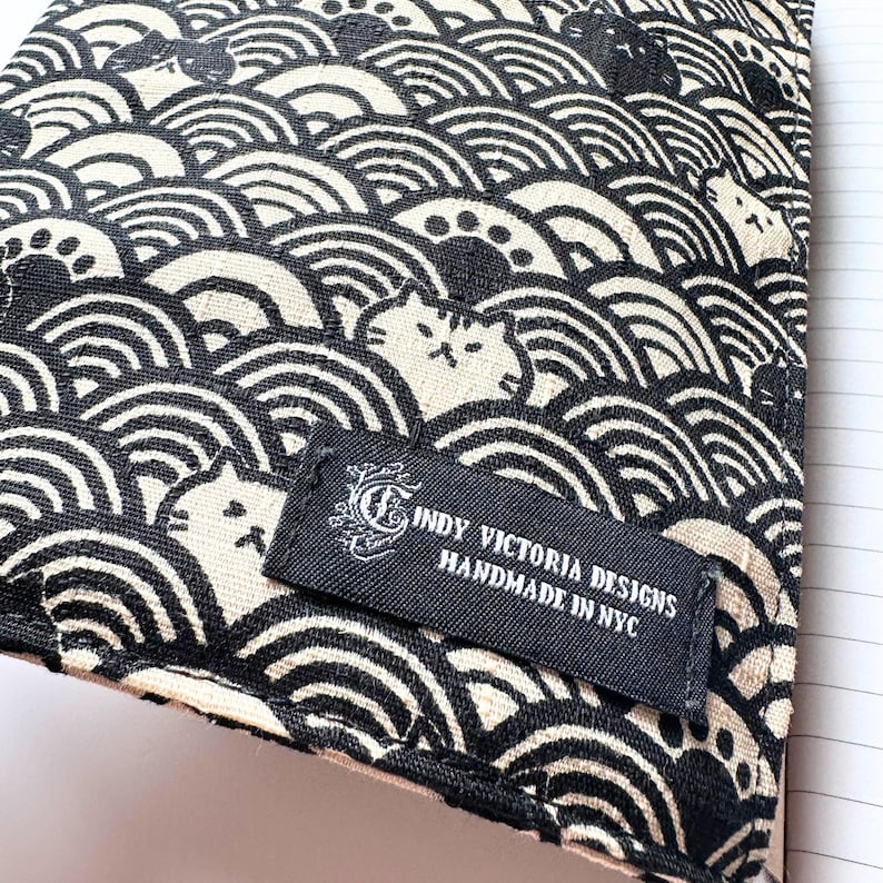 Puede incluir: Tela negra y crema con un patr&oacute;n de olas repetido y caras de gato. Una etiqueta negra dice "INDY VICTORIA DESIGNS HANDMADE IN NYC". La tela probablemente se utiliza para una cubierta de cuaderno o un art&iacute;culo similar.