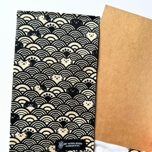 Puede incluir: Funda de tela negra y crema con un patr&oacute;n repetido de olas estilizadas y caras de gato. La funda est&aacute; abierta, revelando una tarjeta marr&oacute;n. El texto "INDY VICTORIA DESIGNS HANDMADE IN NYC" est&aacute; impreso en la funda.