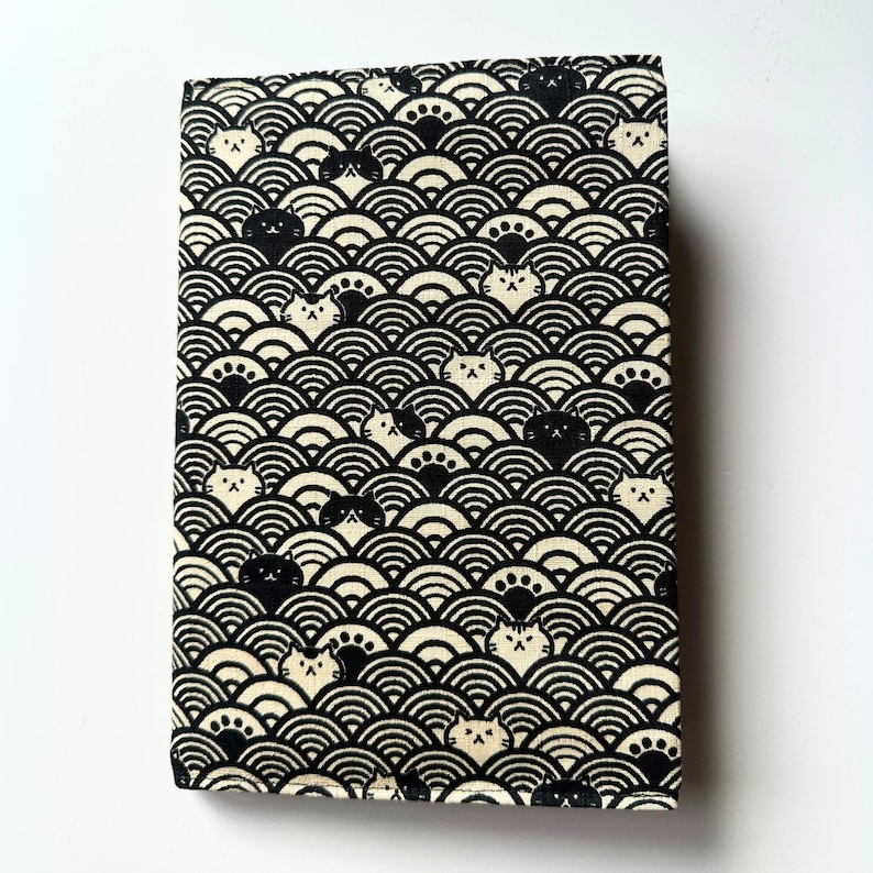 Puede incluir: Un art&iacute;culo rectangular de tela con un patr&oacute;n repetido de formas onduladas en blanco y negro y caras de gato. Los gatos est&aacute;n en varias poses, algunos mostrando sus patas. El art&iacute;culo es probablemente una cubierta de libro o un accesorio similar.
