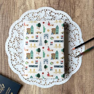 Puede incluir: Un cuaderno con una cubierta de tela blanca con un patrón repetido de símbolos de Nueva York. El diseño incluye edificios, la Estatua de la Libertad, puentes y el texto "I ❤️ NY". El cuaderno está sobre un mantel individual de encaje blanco.