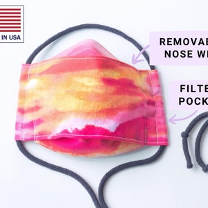Puede incluir: Una mascarilla facial con un vibrante diseño de acuarela en tonos rosa, naranja y amarillo. La mascarilla cuenta con un alambre nasal extraíble y un bolsillo para filtro. La mascarilla tiene presillas grises y está etiquetada como "MADE IN USA".