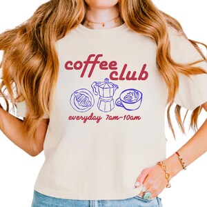 Może przedstawiać: Kremowa koszulka z napisem "coffee club" w bordowym skrypcie. Poniżej niebieskie ilustracje rogalika, kawiarki i filiżanki kawy. Tekst "everyday 7am-10am" znajduje się poniżej.
