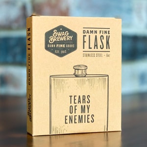 May include: A brown cardboard box with a black and white illustration of a flask. The box has the text "Swag Brewery Damn Fine Goods Est. 2012" and "Damn Fine Flask Stainless Steel - 6oz" printed on it. The flask has the text "Tears of My Enemies" printed on it.