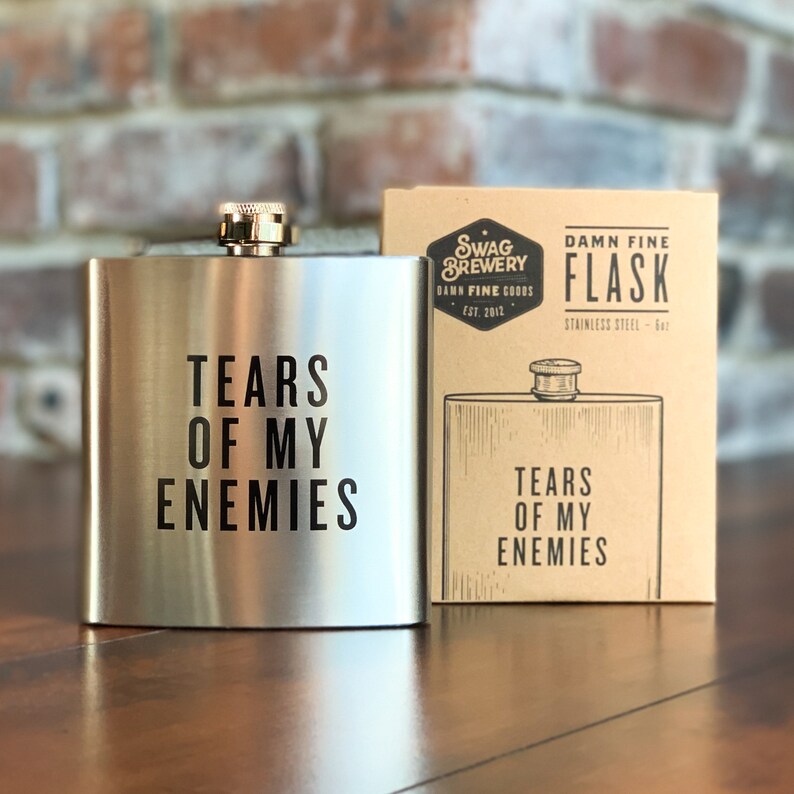 May include: A silver flask with the text "Tears of My Enemies" on it. The flask is next to a brown box with the text "Swag Brewery Damn Fine Coors Est. 2012 Damn Fine Flask Stainless Steel - 8oz" on it.