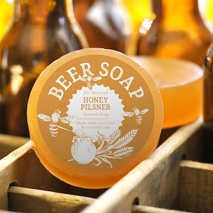 May include: A round bar of honey pilsner scented soap with a white label that reads "BEER SOAP", "All Natural HONEY PILSNER Scented Soap", and "Made with real beer & crushed oats".