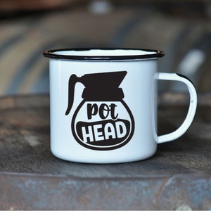 May include: White enamel coffee mug with a black rim and handle. The mug features a black graphic of a coffee pot with the words "pot HEAD" in a white oval. The mug is on a wooden surface.