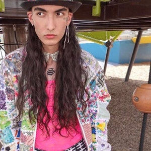 May include: A person wearing a white and multi-colored patterned jacket with a pink tank top underneath. They are also wearing black and white patterned shorts and a black hat with a multi-colored gem design on top.